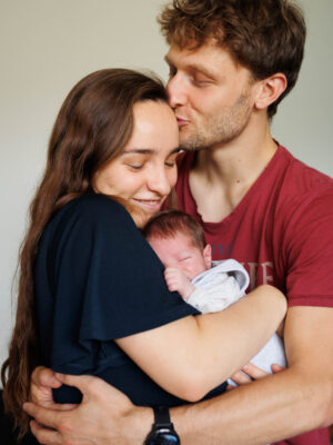 ein junges Elternpaar mit Neugeborenen. Der Mann hält die Frau liebevoll umarmt, in der Mitte das Baby, beide lächeln mit geschlossenen Augen