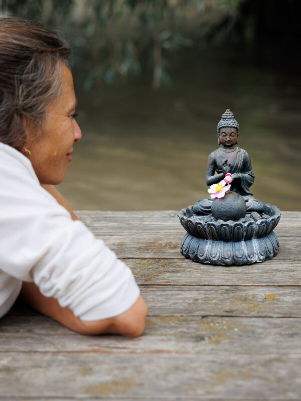 Hebamme Schanti in Teilausschnitt, wie sie am Holz Bootssteg bäuchlings mit aufgestellten Armen liegt und freundlich eine Buddha Figur anschaut