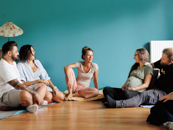 eine Gruppenkurs Situation mit zwei Pärchen, wo die Frauen schwanger sind, sitzen mit der Hebamme am Boden zusammen. Es herrscht eine heiter, interessierte Stimmung, während Hebamme Schanti in der Mitte sitzend, etwas erzählt.