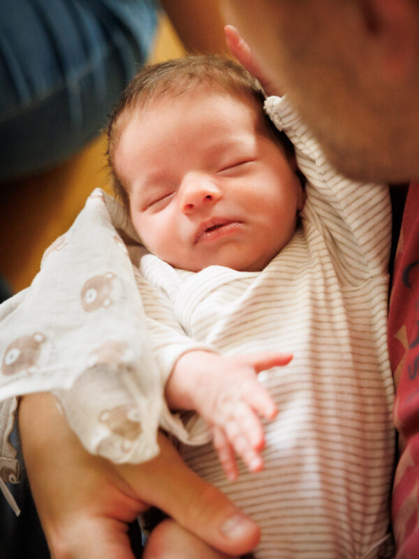 Nahaufnahme eines kleinen Babys, welches in den Armen seines Vaters liegt, mit geschlossenen Augen, die Ärmchen in Bewegung