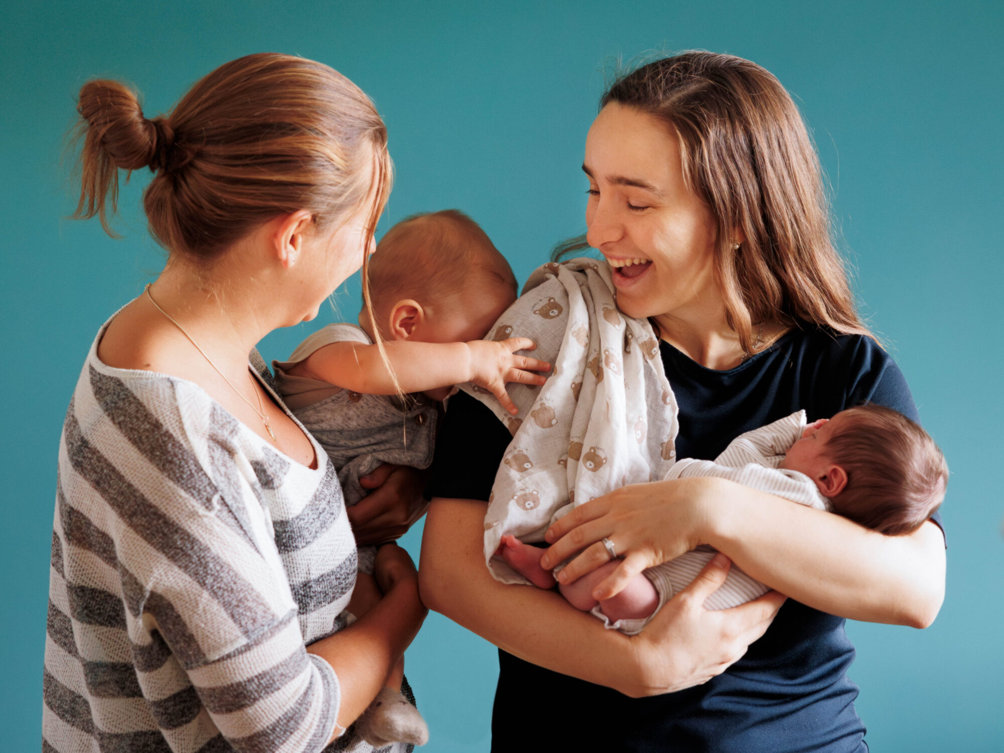 zwei Mütter mit ihren Babys im Arm. Auf der linken Seite trägt die Frau ihr ca. 6 Monate altes Baby auf der Hüfte, welches grade in spielerischer Form den Kopf an den Arm der anderen Frau lehnt. Diese hält ihr Neugeborenes im Wiegegriff und lacht herzlich.