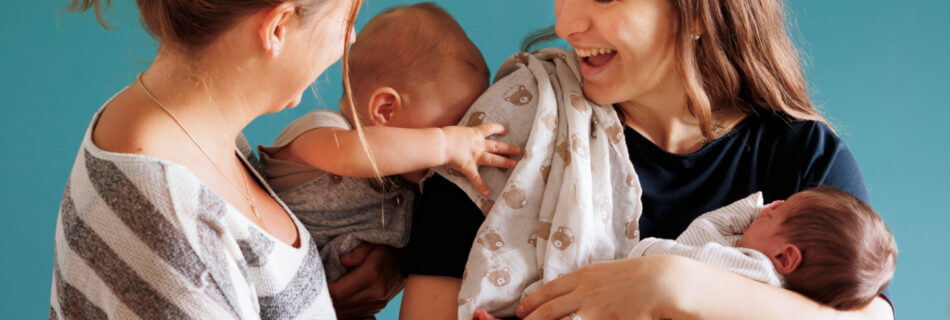 zwei Mütter mit ihren Babys im Arm. Auf der linken Seite trägt die Frau ihr ca. 6 Monate altes Baby auf der Hüfte, welches grade in spielerischer Form den Kopf an den Arm der anderen Frau lehnt. Diese hält ihr Neugeborenes im Wiegegriff und lacht herzlich.
